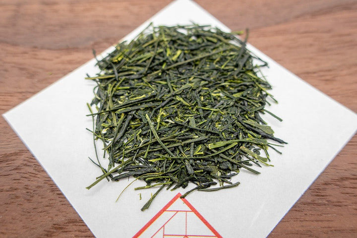 Needle-shaped and deep emerald premium sencha green tea leaves from Yame, Japan on a square white sheet of paper on a wooden plate.