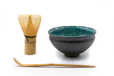 Front side of a matcha bowl, with a dark grey and rough outside and a vibrant and glazed turquoise color on the inside, next to a matcha whisk and a matcha spoon.