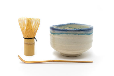 Front of a cream-white matcha bowl with a blue-colored glaze all around its uneven edge, with matcha whisk and a matcha spoon next to it.