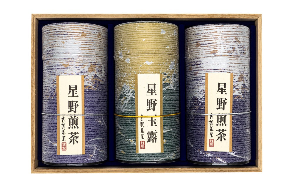 Vue de face de trois boîtes de thé vert en forme de tube violet et doré placées à l'intérieur d'une boîte en bois portant des inscriptions japonaises mentionnant leurs origines (Hoshino sencha et gyokuro).