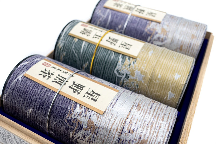 Vue rapprochée de trois boîtes de thé vert en forme de tube violet et doré portant des inscriptions japonaises mentionnant leurs origines (Hoshino sencha et gyokuro).