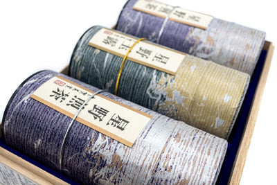 Close view of three purple and golden tube-shaped green tea boxes with Japanese markings on them mentioning their origins (Hoshino sencha and gyokuro).