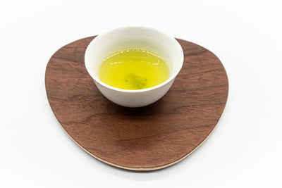 Side view of a small white porcelain cup filled with brewed premium Japanese sencha green tea on a triangular-shaped wooden plate.