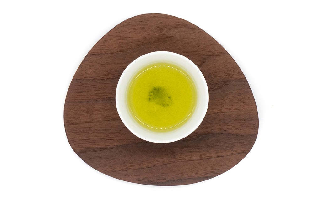 Vue de dessus d'une petite tasse en porcelaine blanche remplie de thé vert sencha japonais infusé de première qualité sur une assiette en bois de forme triangulaire.
