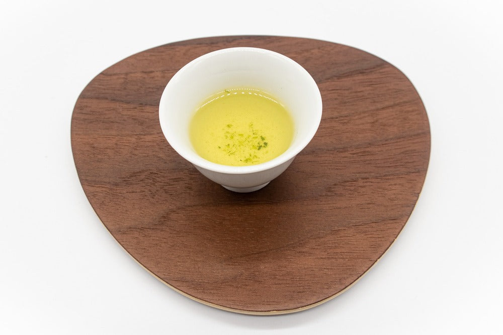 Vue latérale d'une petite tasse en porcelaine blanche remplie de thé vert dento hon gyokuro infusé sur une assiette en bois de forme triangulaire.