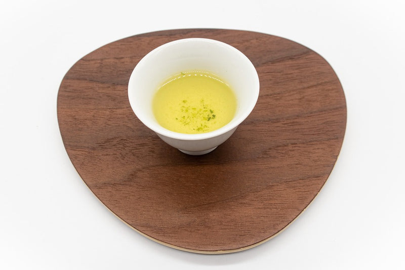 Side view of a small white porcelain cup filled with brewed dento hon gyokuro green tea on a triangular-shaped wooden plate.
