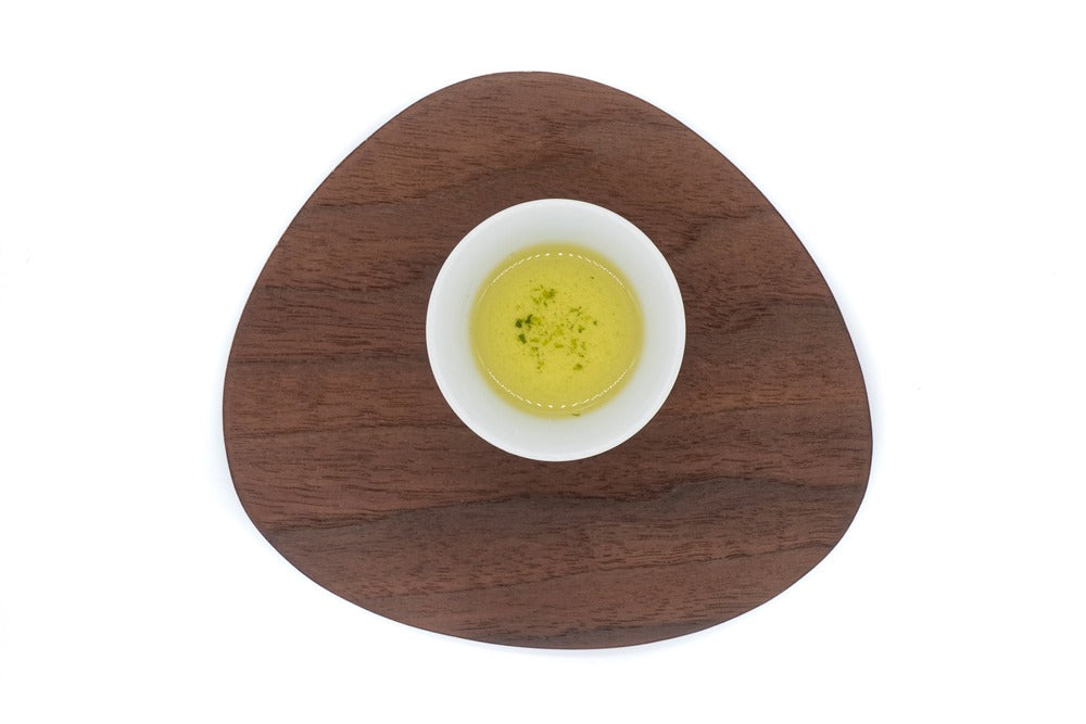 Vue de dessus d'une petite tasse en porcelaine blanche remplie de thé vert dento hon gyokuro infusé sur une assiette en bois de forme triangulaire.