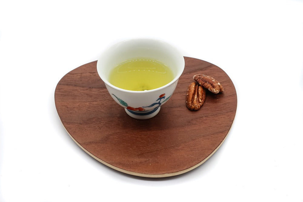 Tasse en porcelaine blanche remplie de thé blanc japonais de qualité supérieure infusé, placée sur une assiette en bois de forme triangulaire, avec deux noix grillées à côté.