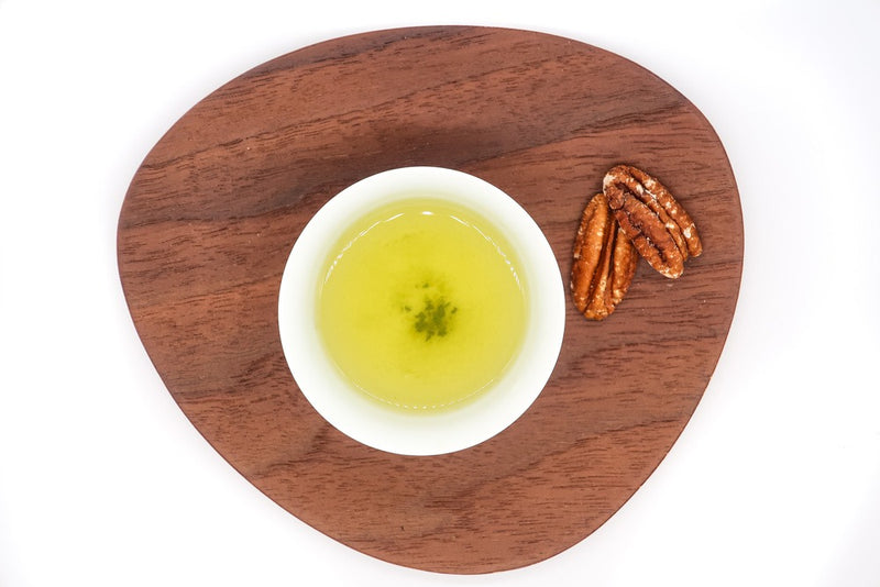 Top view of a small white porcelain cup filled with brewed premium Japanese white tea on a triangular-shaped wooden plate, with two roasted nuts next to it.