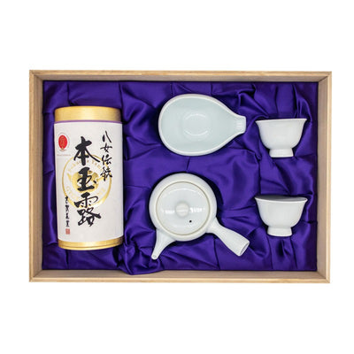 Open wooden box containing a set made of a tube-shaped golden box with a white paper label wrapped around it with hand-written Japanese calligraphy, a white porcelain tea pot, a white porcelain water cooler and two small white porcelain tea cups resting a