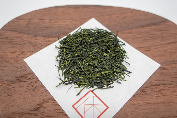 Needle-shaped and deep emerald premium sencha green tea leaves from Yame, Japan on a square white sheet of paper on a wooden plate.