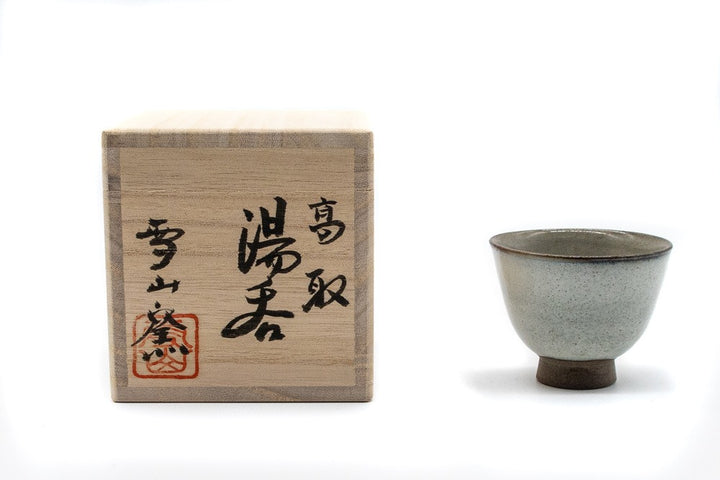 Face avant d'une petite et délicate tasse grise faite à la main pour boire du thé vert gyokuro, fabriquée à Koishiwara, Fukuoka, Japon par Onimaru le Second, à côté d'une boîte en bois avec une calligraphie japonaise à la main qui accompagne la tasse.